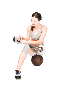 smiling disabled sportswoman with prosthesis sitting on basketball ball and tying shoelaces isolated on white