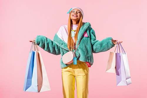 happy, stylish african american woman standing with closed eyes and holding shopping bags isolated on pink, fashion doll concept