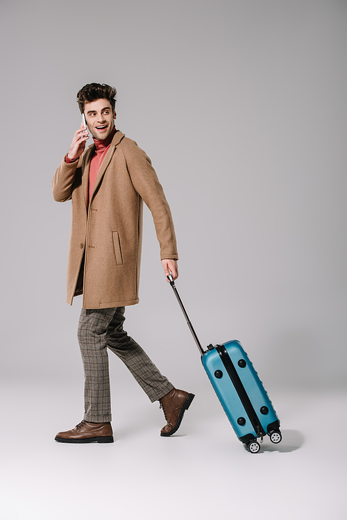 smiling man in beige coat talking on smartphone and walking with travel bag on grey