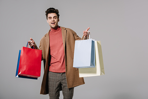 cheerful stylish man in beige coat holding shopping bags, isolated on grey