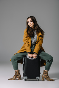 attractive woman in beige coat sitting on travel bag on grey