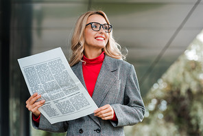 attractive businesswoman in coat and glasses smiling and holding newspaper