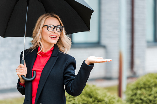 attractive and smiling businesswoman in black coat with outstretched hand holding umbrella