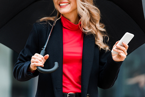 cropped view of blonde businesswoman in black coat smiling and holding smartphone