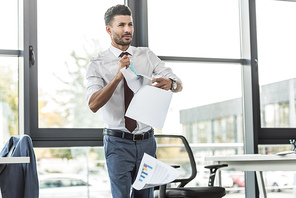 displeased businessman tearing documents and looking away while standing near window in office