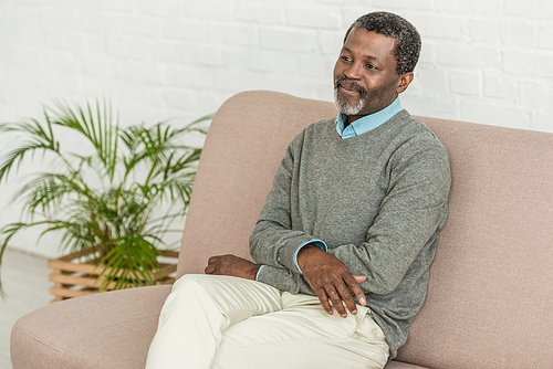 dreamy, smiling african american man sitting on sofa at home