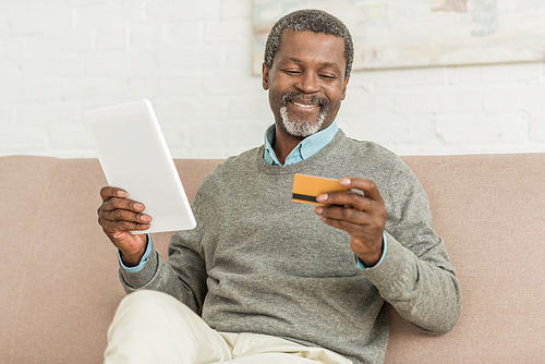 smiling african american man holding digital tablet and credit card while sitting on sofa