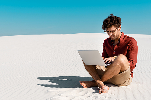 young freelancer on sandy beach using laptop against clear blue sky