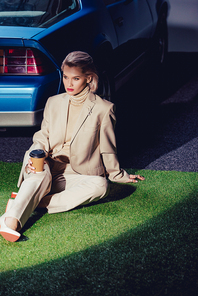 attractive and stylish woman in suit sitting near retro car and holding paper cup