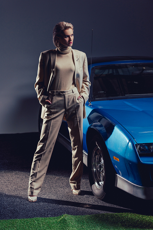 attractive and stylish woman in suit with hands in pockets standing near retro car