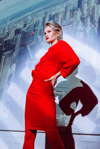 low angle view of attractive and stylish woman with hand on hip in red dress on city background