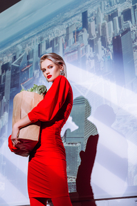 low angle view of attractive and stylish woman in red dress holding paper bag with food on city background