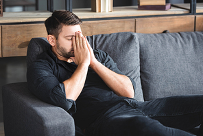 man with headache sitting on sofa and obscuring face in apartment