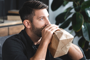 handsome man with panic attack breathing in paper bag in apartment