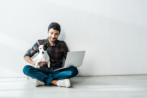 handsome and smiling bi-racial man with laptop holding jack russell terrier