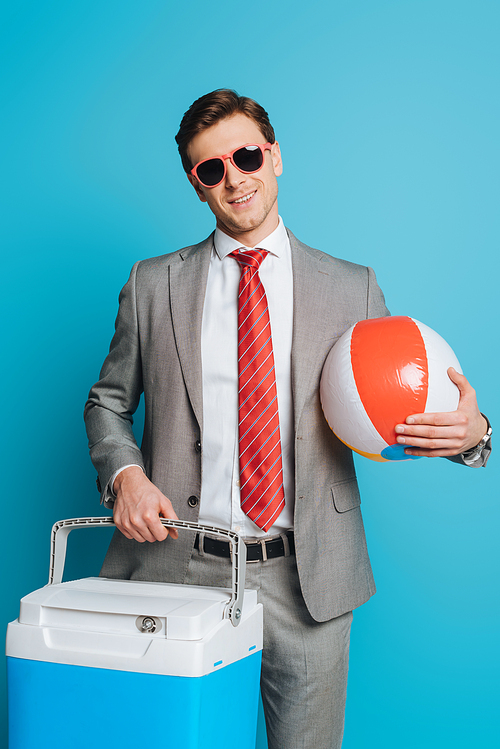 smiling businessman in sunglasses holding portable fridge and inflatable ball on blue background