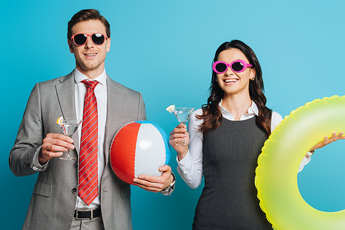 two cheerful businesspeople with glasses of cocktail, inflatable ball and swim ring  on blue background