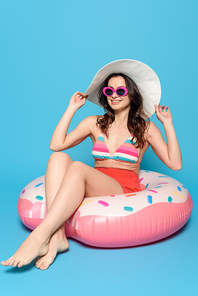cheerful, stylish woman in sunglasses touching sun hat while sitting on swim ring on blue background
