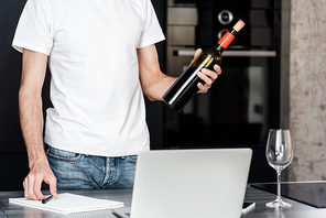 Cropped view of man holding wine bottle near laptop and notebook on worktop in kitchen