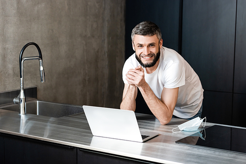 Handsome freelancer smiling at camera near medical mask and laptop on kitchen worktop