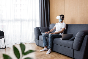 Selective focus of man in vr headset and medical mask sitting near laptop on couch in living room