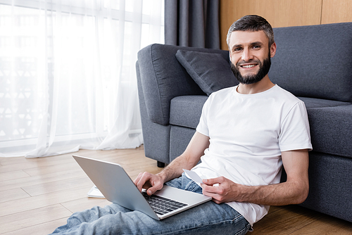 Handsome man smiling at camera while using laptop and credit card on floor at home