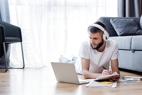 Man in headphones writing on notebook and looking at laptop during webinar in living room