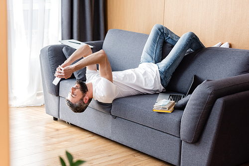 Side view of man using smartphone near books and laptop on sofa at home