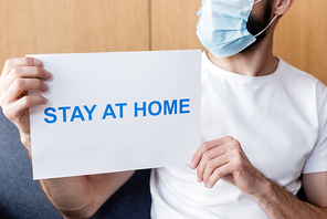 Cropped view of man in medical mask holding card with stay at home lettering on couch