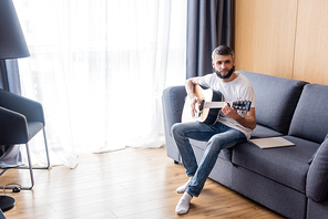 Handsome man playing acoustic guitar near laptop on couch at home