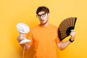 displeased man holding electric and hand fans while suffering from heat on yellow