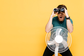 shocked man looking through binoculars while standing with electric fan on yellow