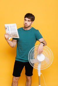 young man suffering from heat with electric fan and newspaper as hand fan on yellow