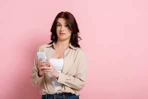Confused woman using smartphone and looking at camera on pink background