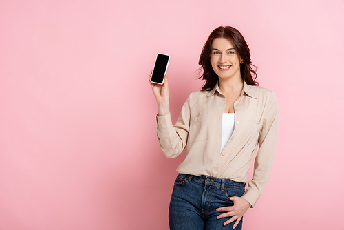 Beautiful brunette woman smiling at camera while holding smartphone on pink background