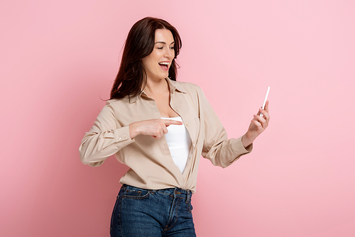 Positive woman pointing with finger at smartphone on pink background