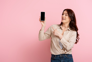 Happy brunette woman pointing with finger at smartphone with blank screen on pink background