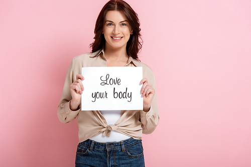 Beautiful woman smiling and showing card with love your body lettering on pink background, concept of body positive