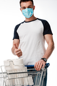 man in medical mask showing thumb up near shopping cart with toilet paper isolated on white
