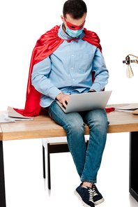 freelancer in medical mask and superhero cape using laptop and sitting on table isolated on white