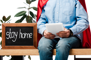 cropped view of man in superhero cape using digital tablet near chalk board with stay home lettering isolated on white