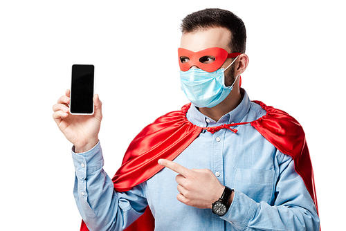 man in superhero costume and medical mask pointing with finger at smartphone with blank screen isolated on white