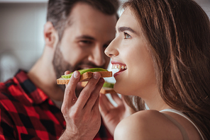 selective focus of smiling man feeding happy girlfriend with fresh avocado toast
