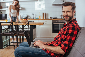 selective focus of handsome freelancer smiling at camera while working on laptop in kitchen near girlfriend on background