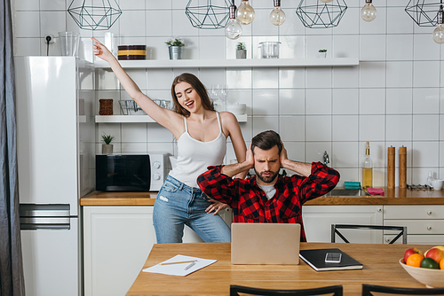 cheerful girl fooling around and dancings near boyfriend covering ears with hands while sitting near laptop and papers