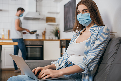 selective focus of freelancer in medical mask working on laptop in kitchen near boyfriend on background