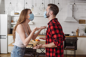 young couple in medical masks quarreling and gesturing in kitchen