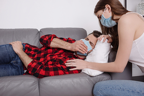 young woman in medical mask squatting near ill boyfriend lying on sofa in medical mask and sneezing