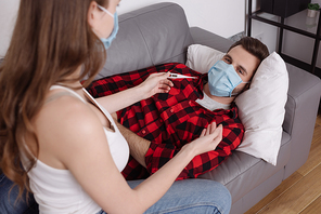 selective focus of girl holding thermometer near diseased boyfriend lying on sofa in medical mask