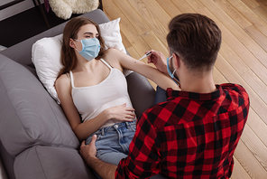 overhead view of man looking at thermometer near diseased girlfriend in medical mask lying on sofa